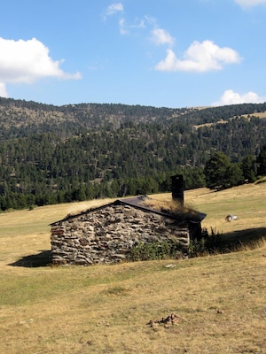 Una casa de piedra con techo de paja en un campo de hierba, rodeada por una cadena montañosa boscosa.