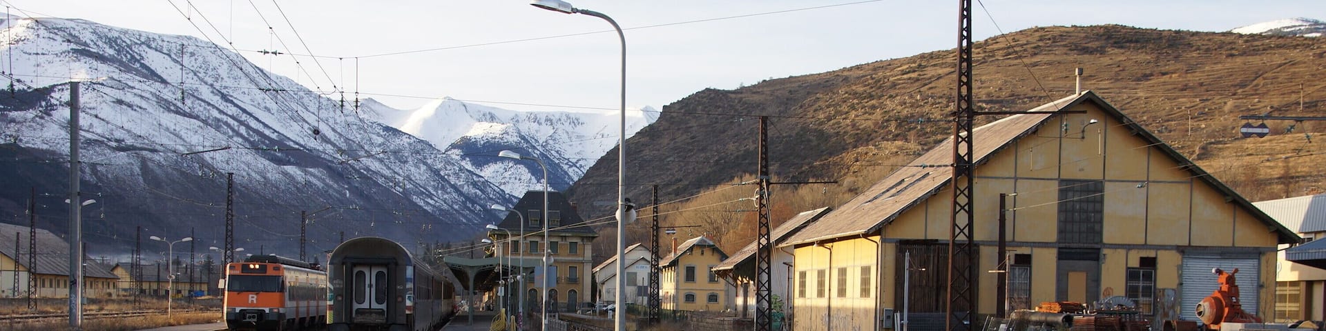A railway station with a train on the tracks, a platform, and a mountainous backdrop.