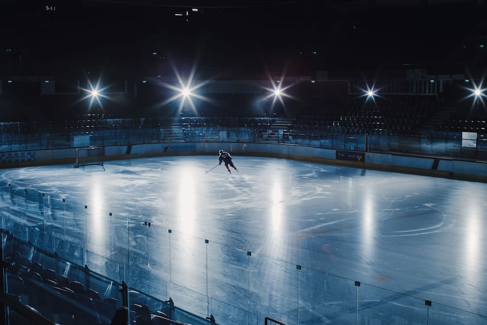 Una pista de hielo cubierta con un jugador en movimiento, luces brillantes en el techo y gradas de espectadores vacías.