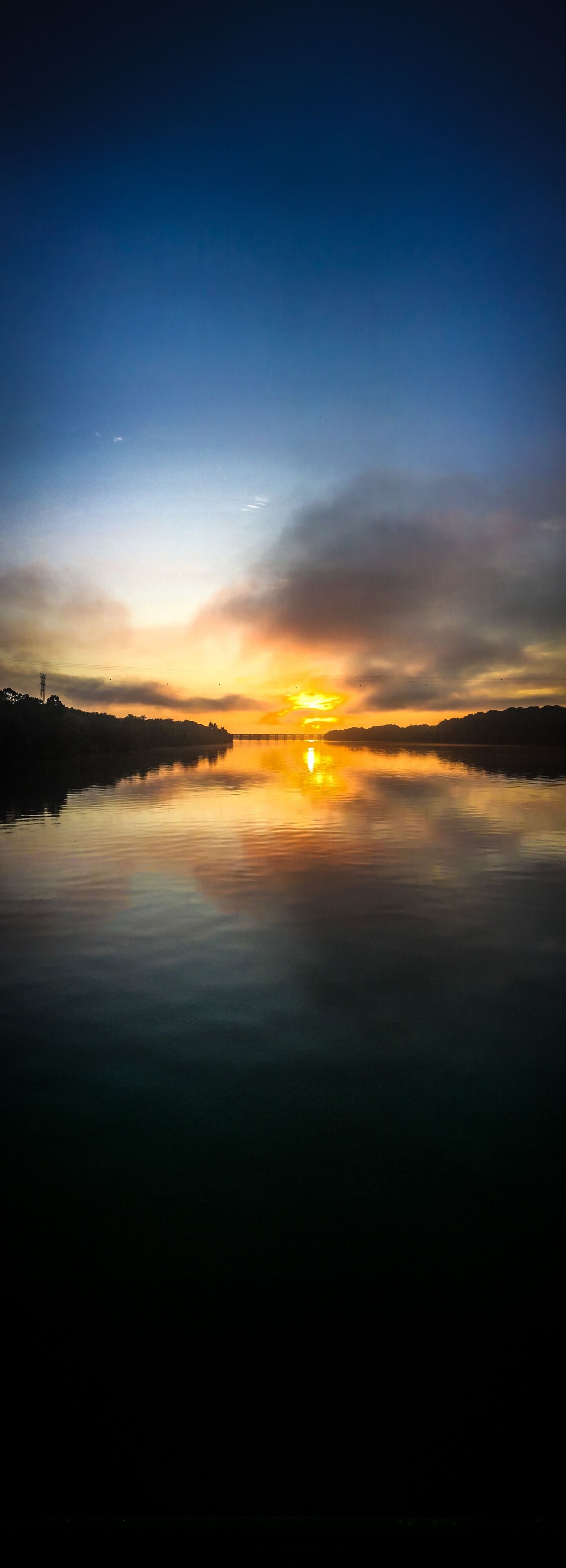 A calm river reflecting the sunset with a silhouette of trees and a distant power line structure.
