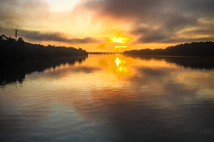 Un río tranquilo reflejando la puesta de sol, con el perfil oscuro de árboles y una estructura de líneas eléctricas a lo lejos.
