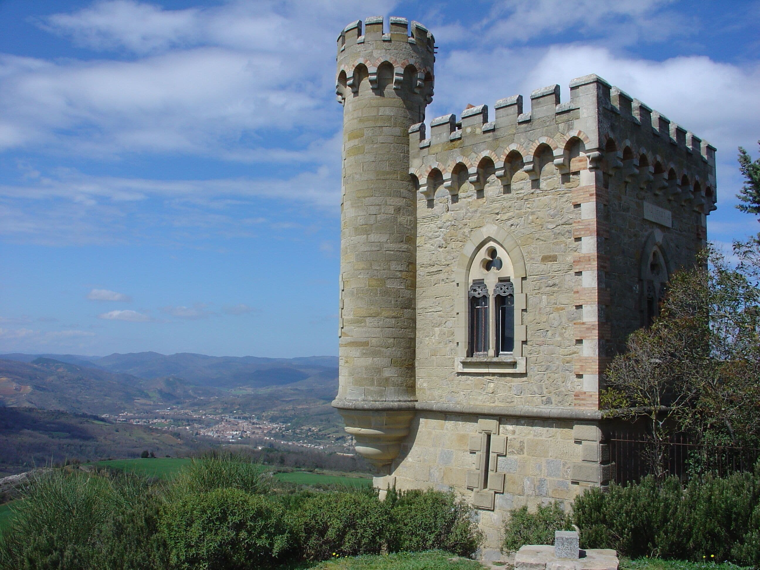 A medieval stone tower with a battlement, arched window, and a view of a valley with buildings in the distance.