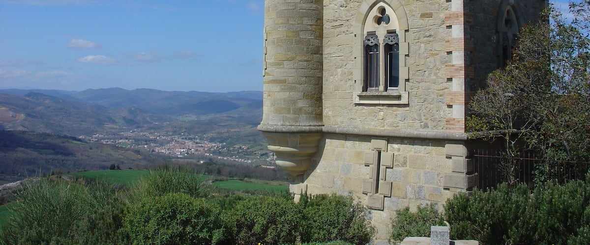 A medieval stone tower with a battlement, arched window, and a view of a valley with buildings in the distance.
