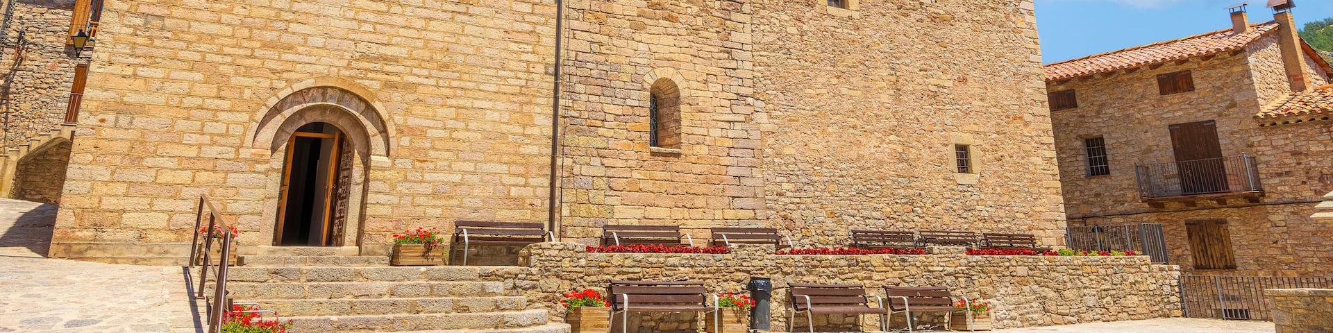 A historic stone building with a bell tower, steps leading to the entrance, and benches with flower boxes.