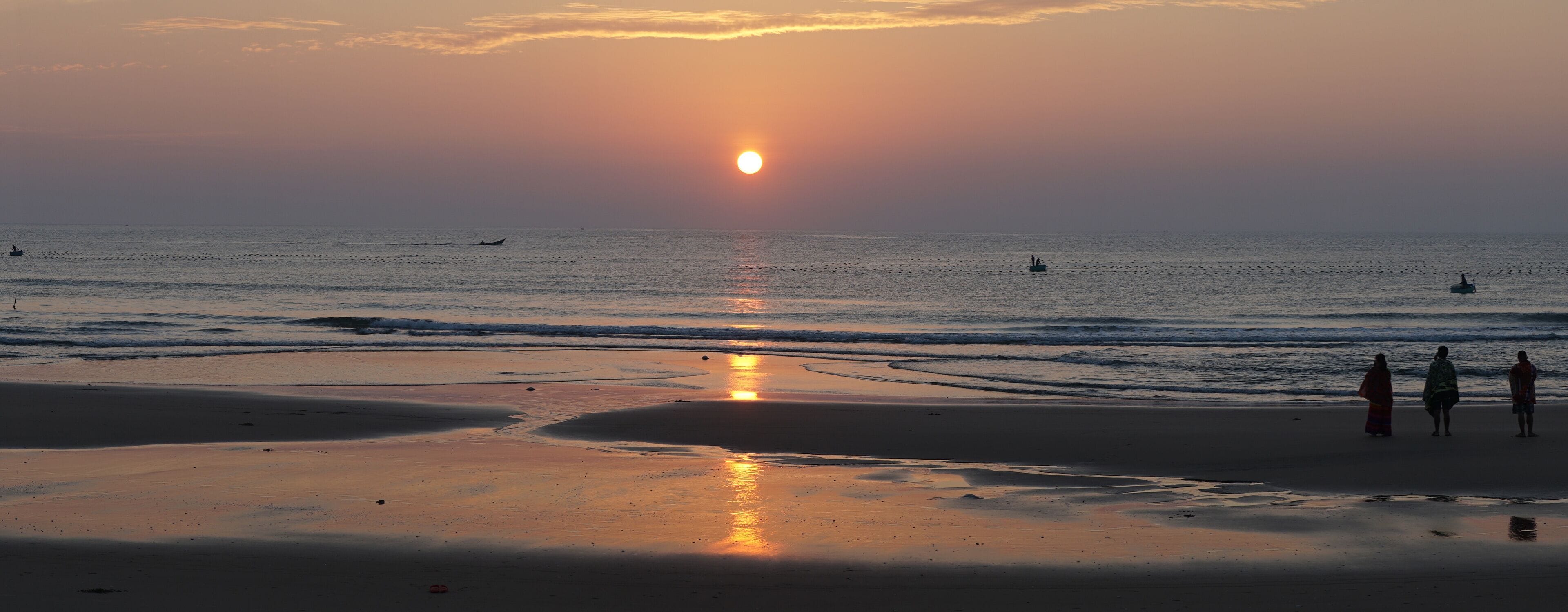 A beach at sunset with the sun low on the horizon, calm sea, and a few boats.