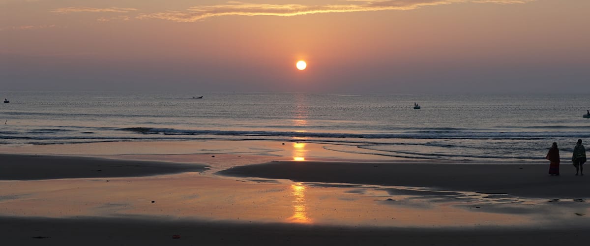 A beach at sunset with the sun low on the horizon, calm sea, and a few boats.