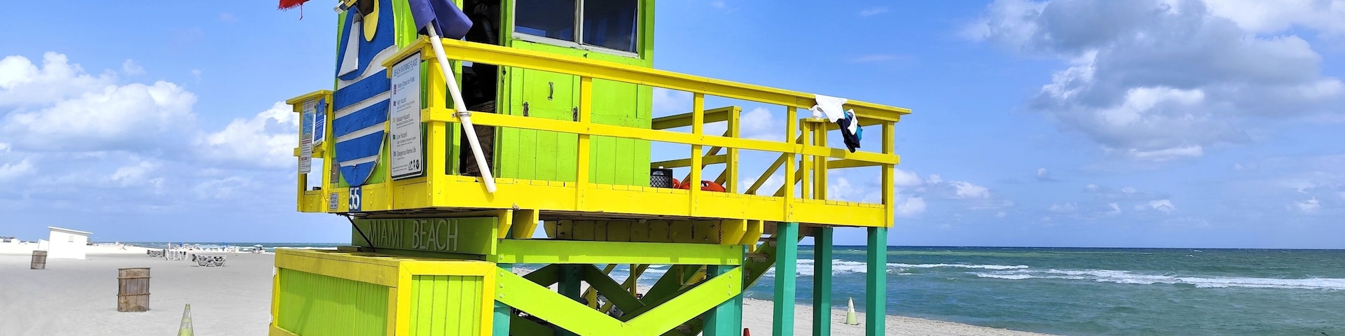 Uma torre de salva-vidas com uma bandeira vermelha no topo, situada numa praia arenosa com o oceano ao fundo.