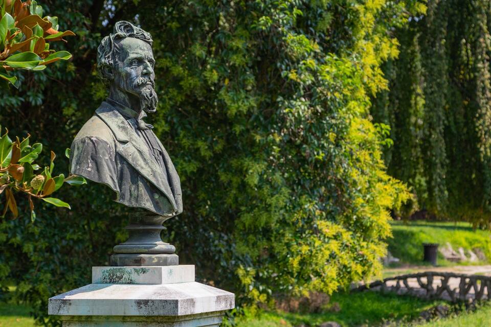 Une statue dans un parc, entourée d’une végétation luxuriante, avec un pont de pierre en arrière-plan.
