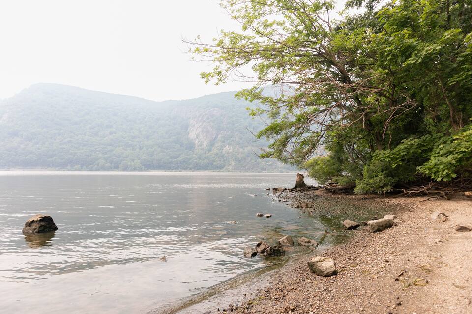 A calm lake with a rocky shoreline, surrounded by dense green trees and mountains in the background.