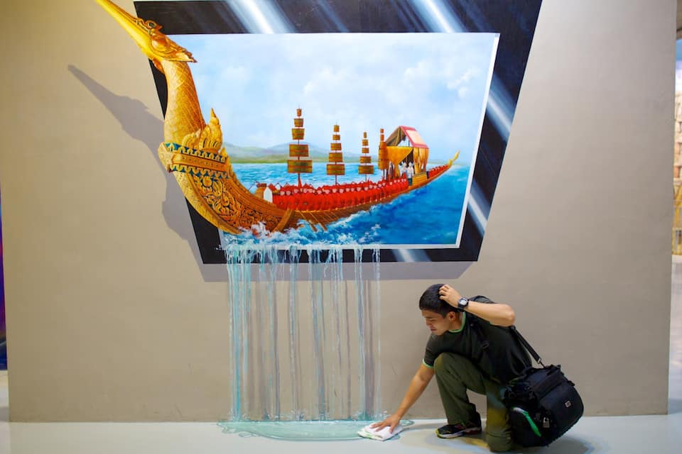 A large painting of a golden ship on water, a person crouching in front of it, and a wall with a partial view of a blue and purple abstract design.