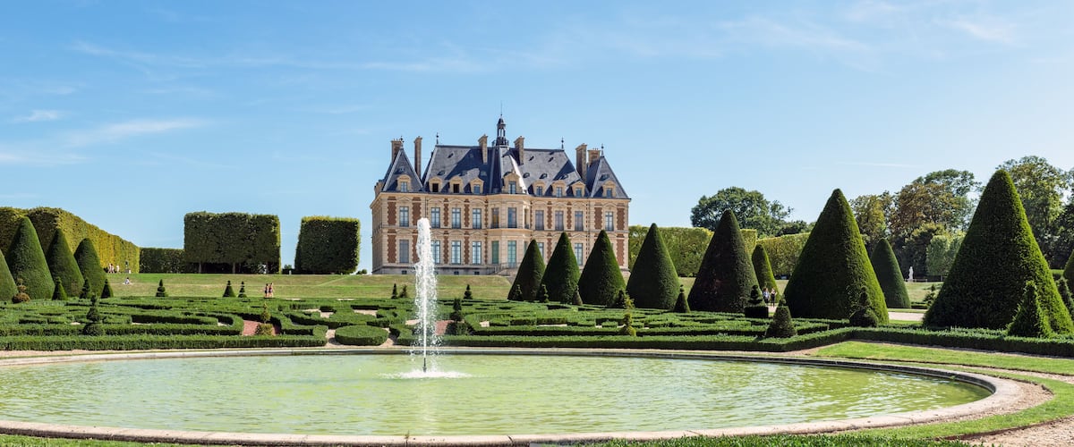 A formal garden with a fountain, neatly trimmed hedges, and a large building in the background.