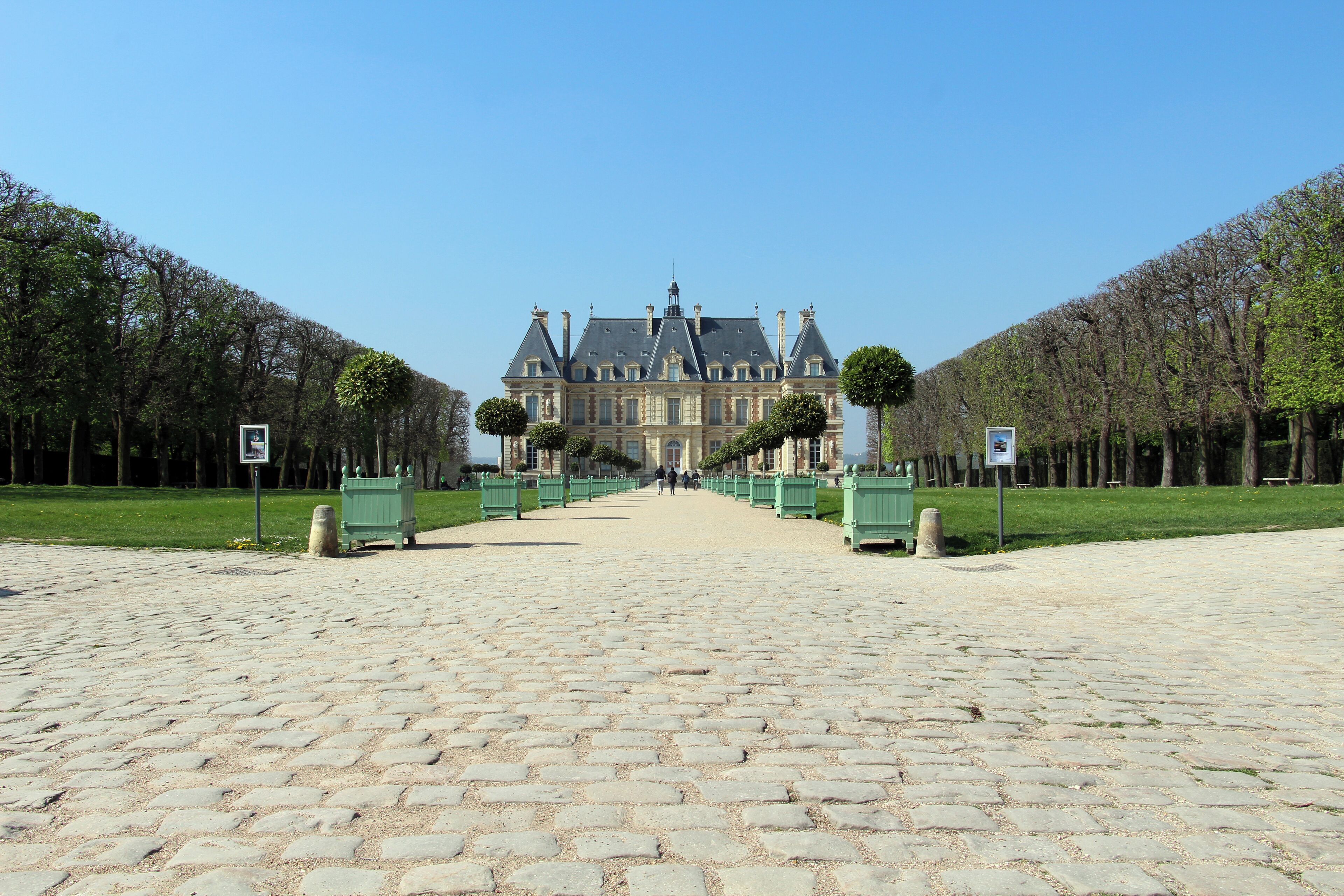A cobblestone pathway leading to a grand château with symmetrical gardens and a clear blue sky.