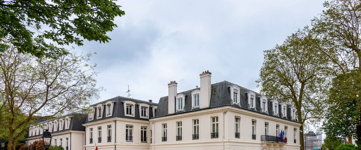 A large, historic white building with multiple chimneys and balconies, surrounded by trees and a gated entrance.