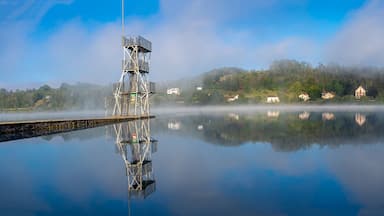 A calm lake with a metal structure on the left, surrounded by mist-covered hills and houses.