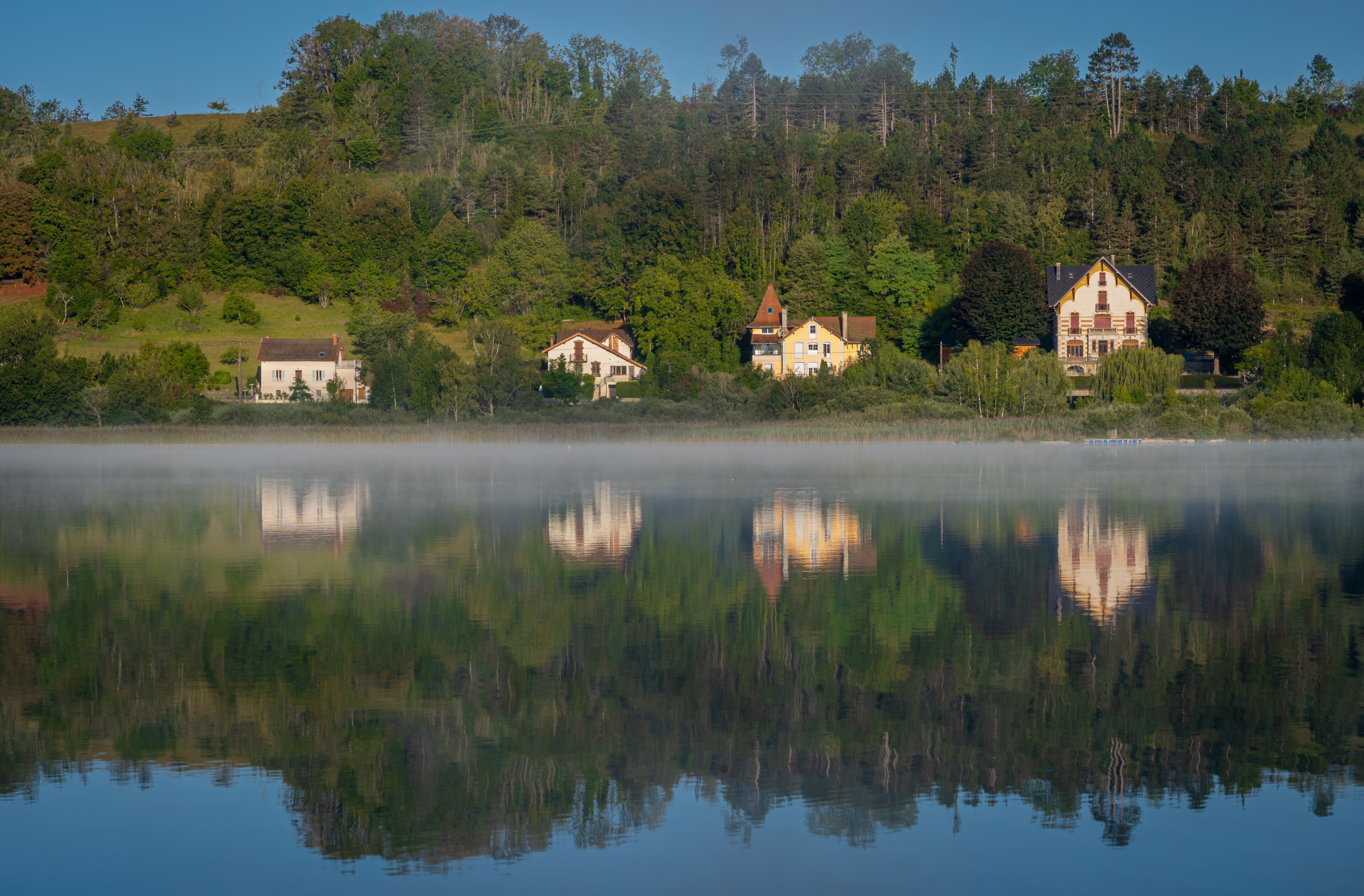 A calm lake with reflections of houses and trees, surrounded by a forested hillside.