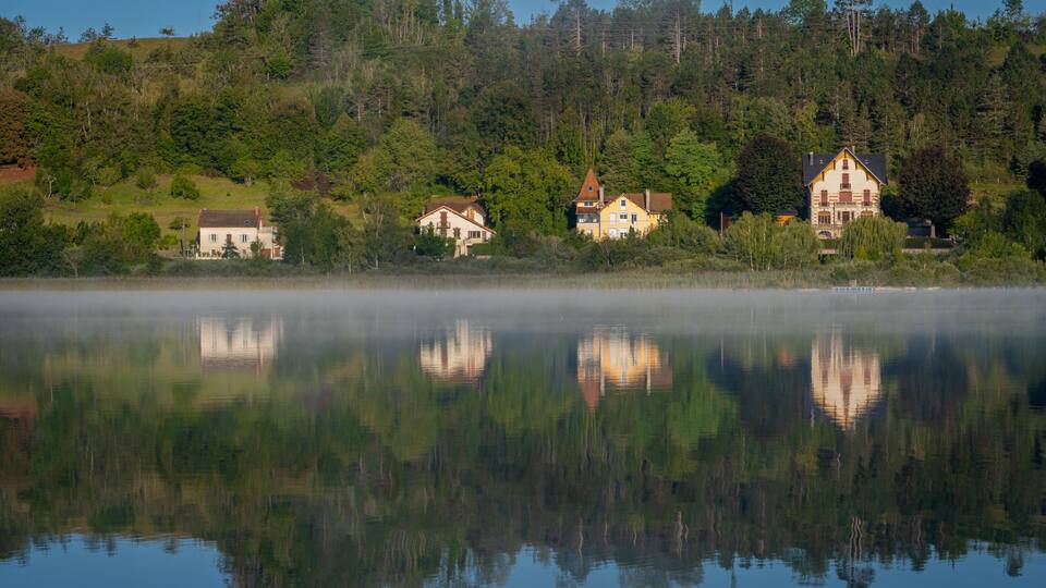 A calm lake with reflections of houses and trees, surrounded by a forested hillside.