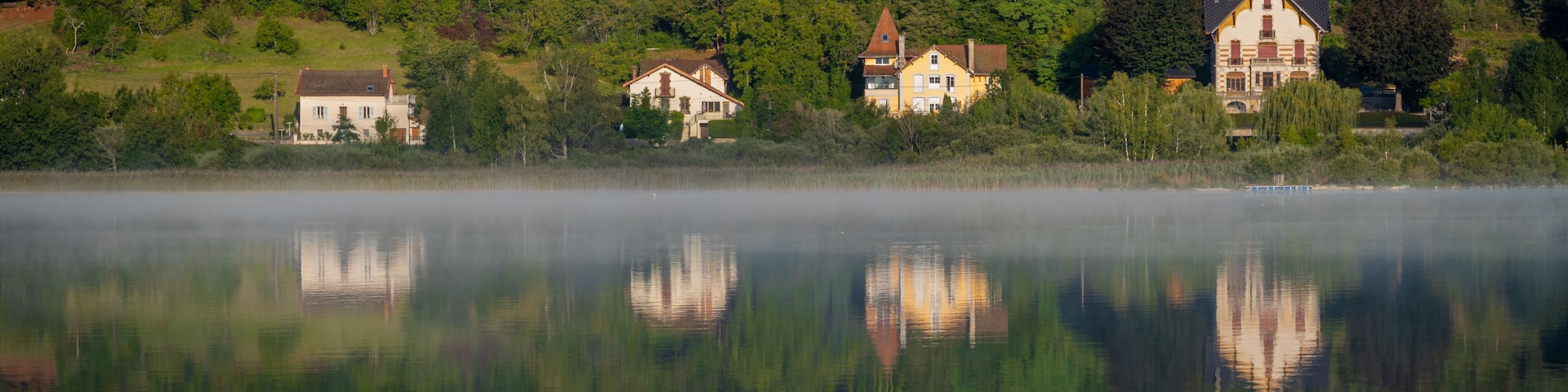 Un lago tranquilo con reflejos de casas y árboles, rodeado de una ladera boscosa.