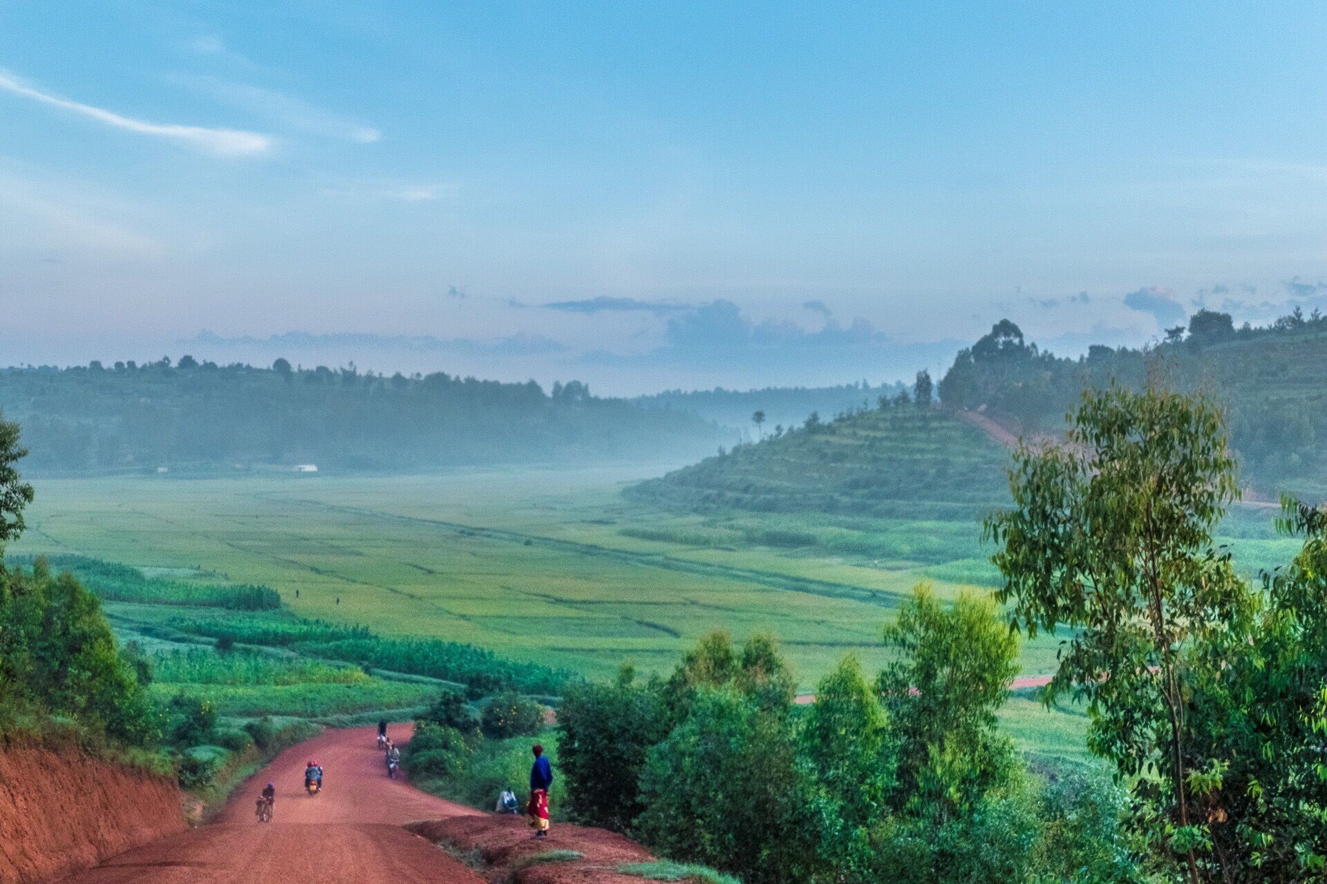 A rural landscape with terraced fields, a dirt road, and a few people riding motorcycles.