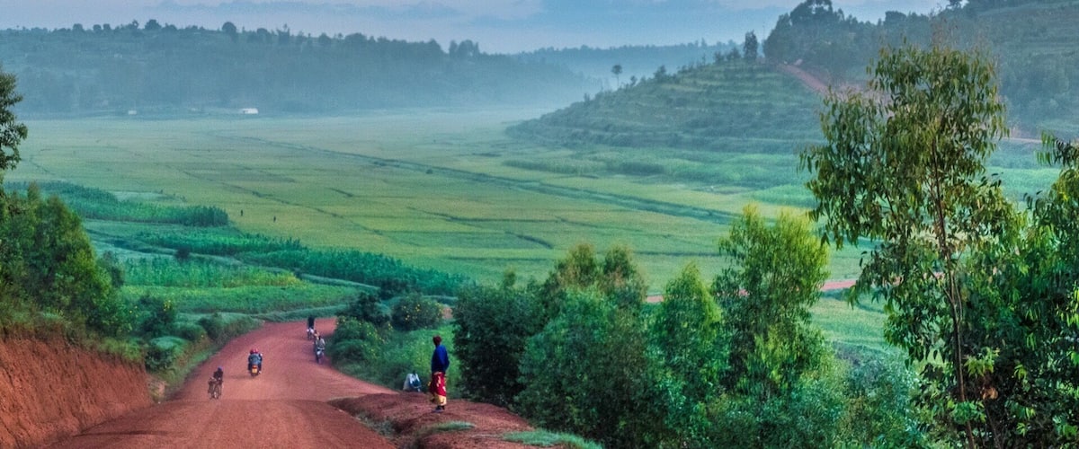 A rural landscape with terraced fields, a dirt road, and a few people riding motorcycles.