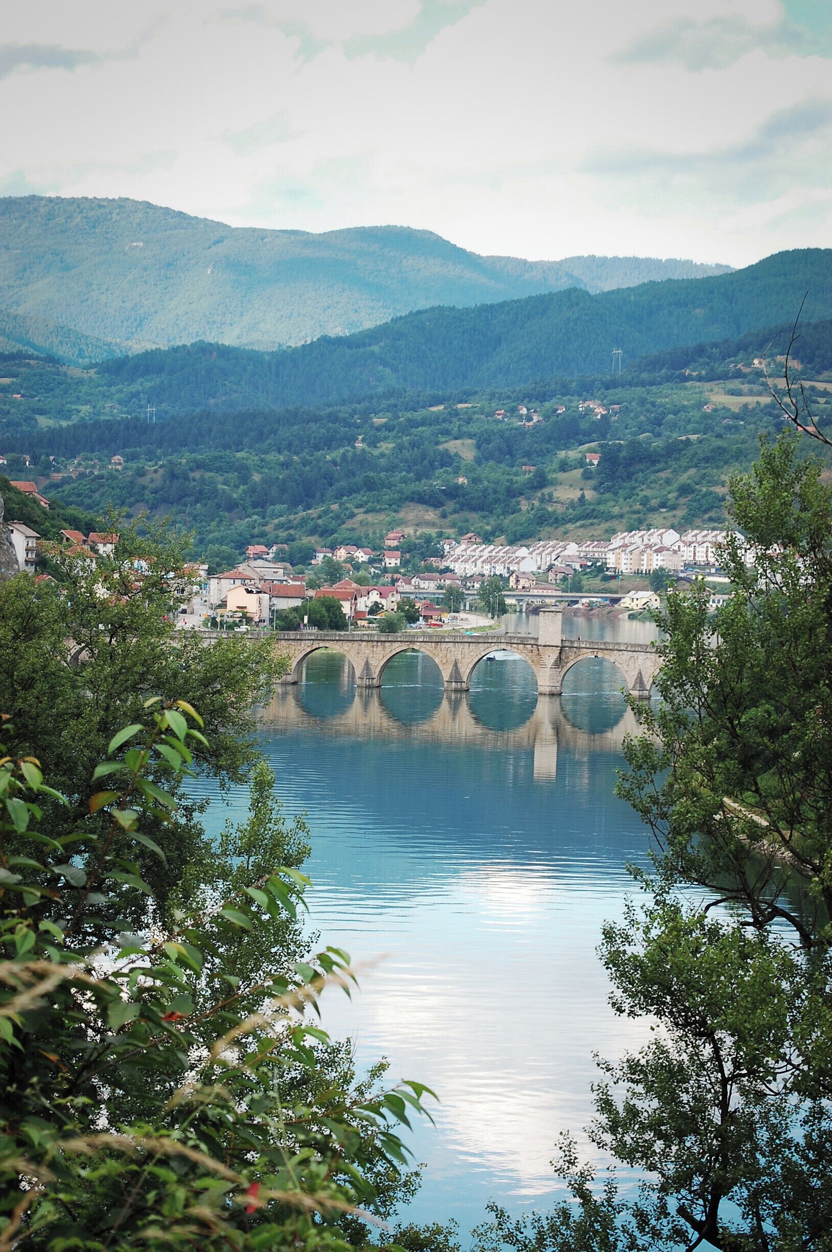 A river with a stone bridge, surrounded by green hills and houses.