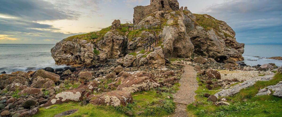 A rocky coastal landscape with a ruined stone structure and a gravel path leading to it.