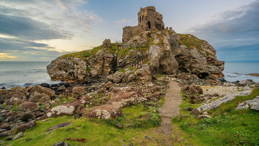 Un paisaje costero rocoso con una estructura de piedra en ruinas y un camino de grava que la atraviesa.