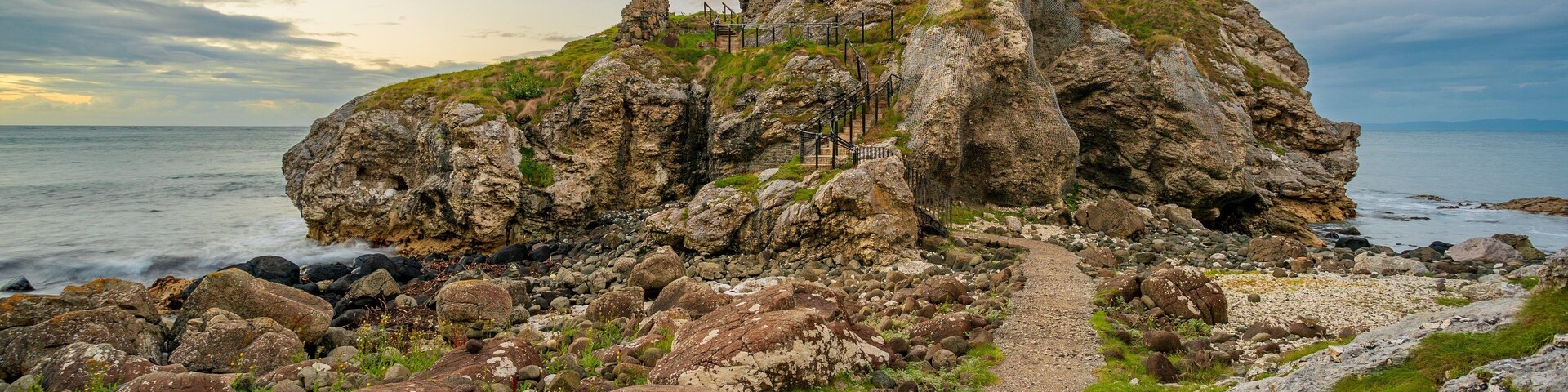 Un paisaje costero rocoso con una estructura de piedra en ruinas y un camino de grava que la atraviesa.