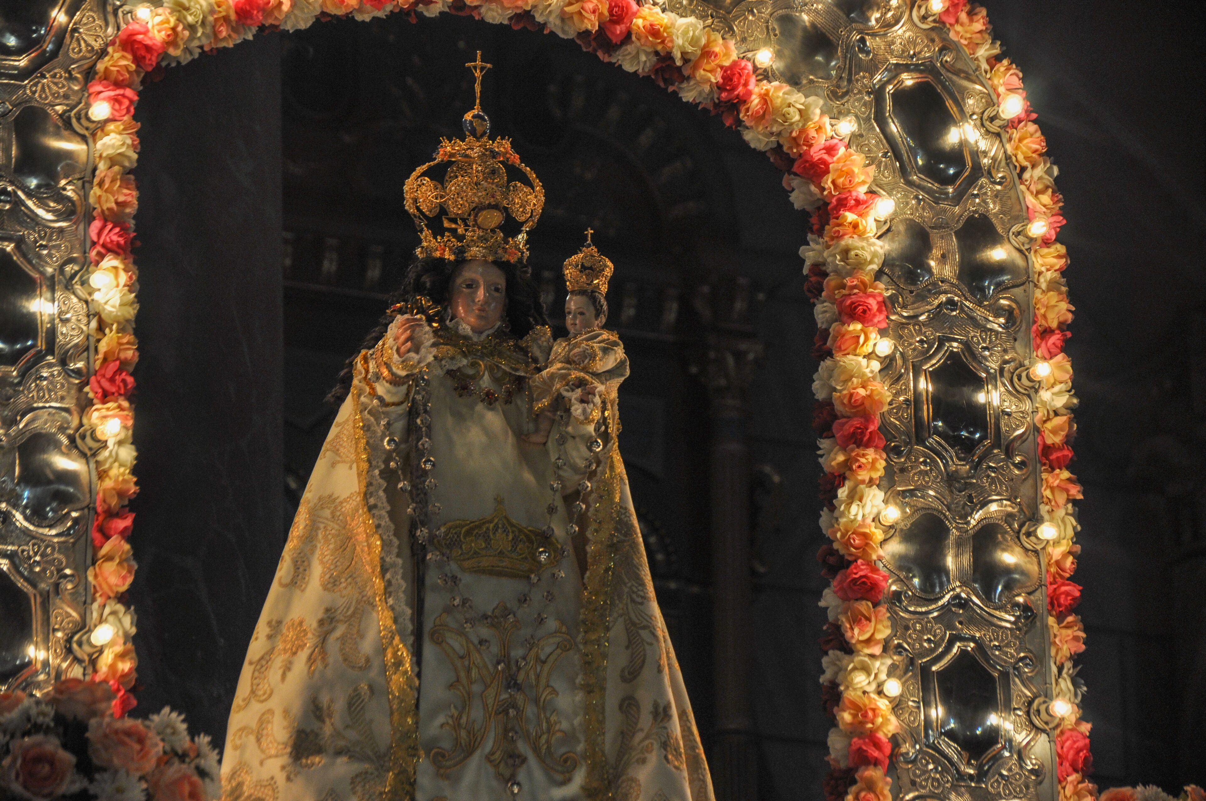 A person in elaborate golden robes and a crown, standing under an ornate arch decorated with flowers.
