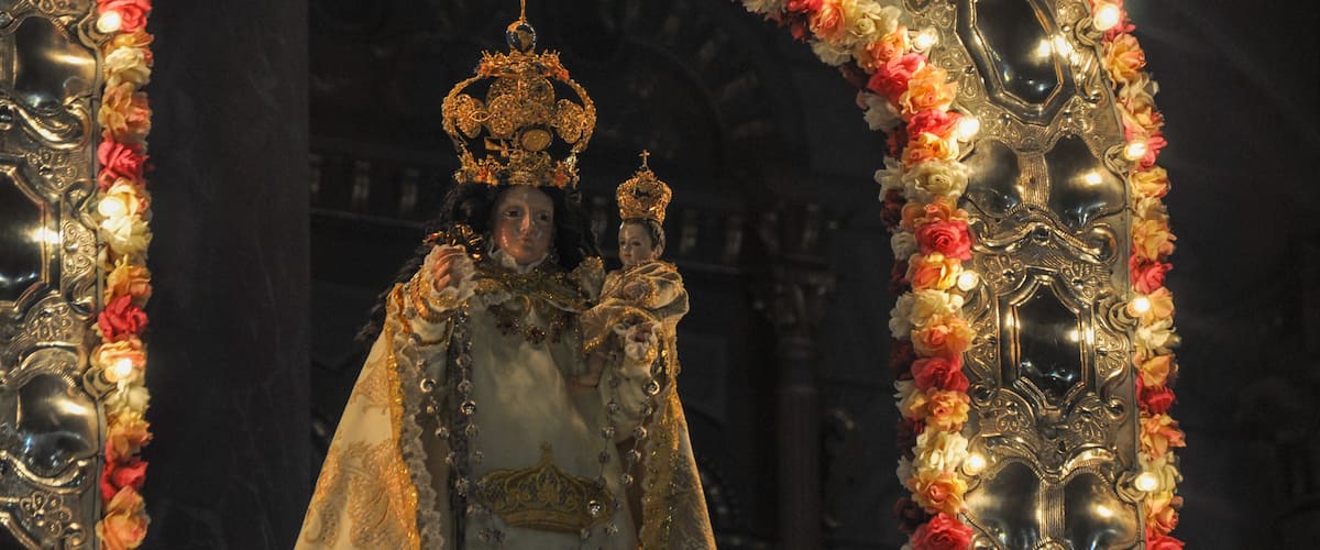 A person in elaborate golden robes and a crown, standing under an ornate arch decorated with flowers.