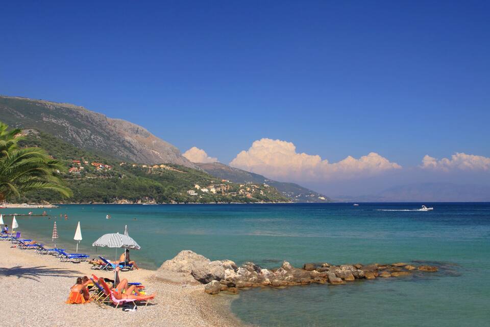 A beach with clear turquoise water, rocky shoreline, and a mountain in the background.