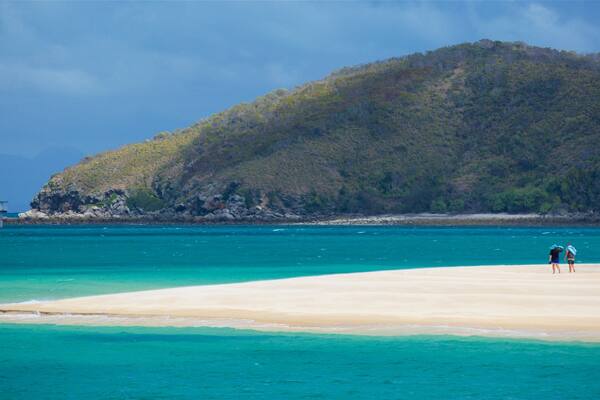 A beach with clear turquoise water, a sandy shore, and a forested hill in the background.