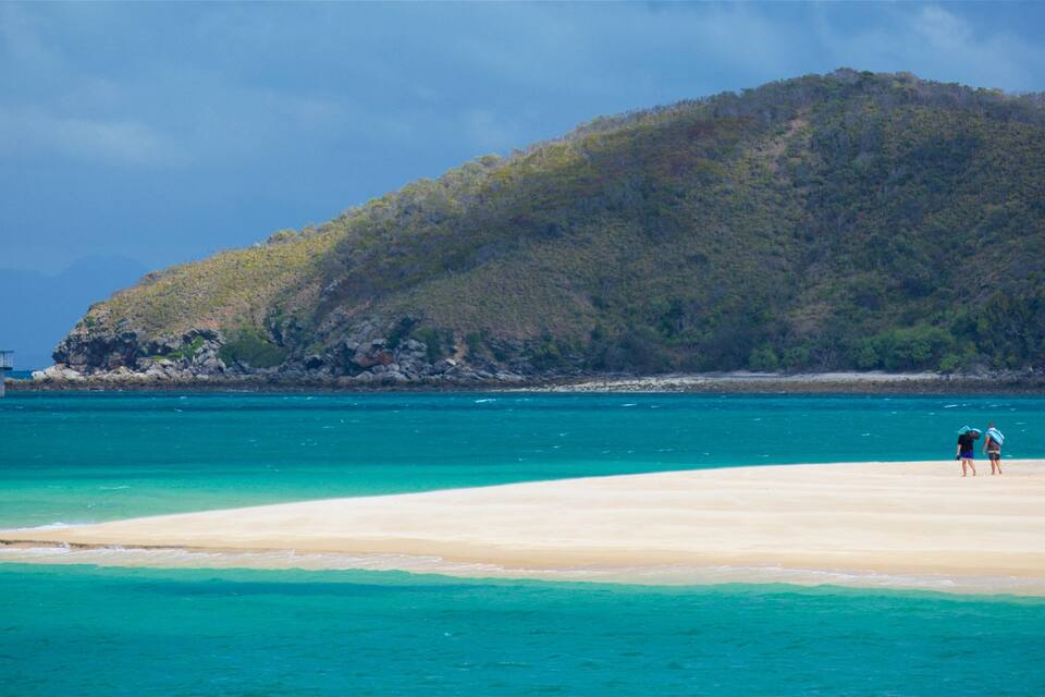 A beach with clear turquoise water, a sandy shore, and a forested hill in the background.