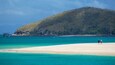 A beach with clear turquoise water, a sandy shore, and a forested hill in the background.