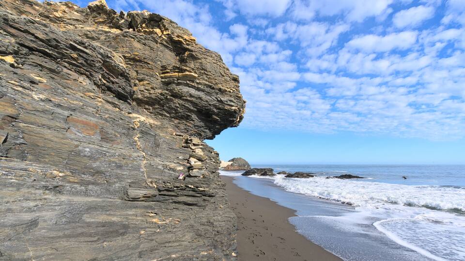 A rocky cliff with layered rock formations and a sandy beach with waves crashing onto the shore.