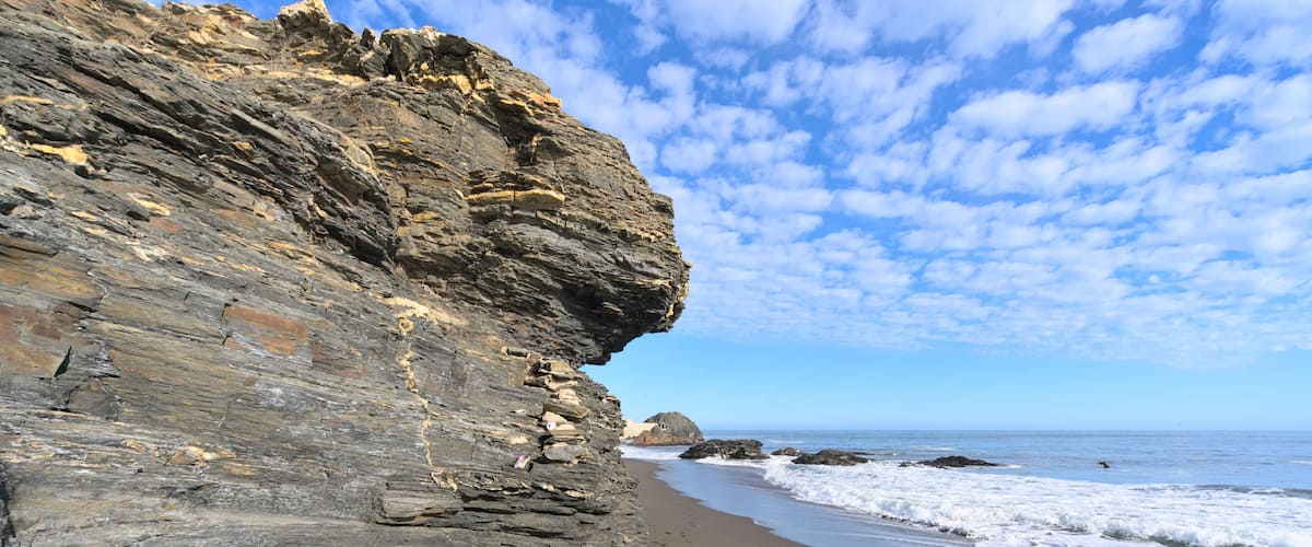 A rocky cliff with layered rock formations and a sandy beach with waves crashing onto the shore.