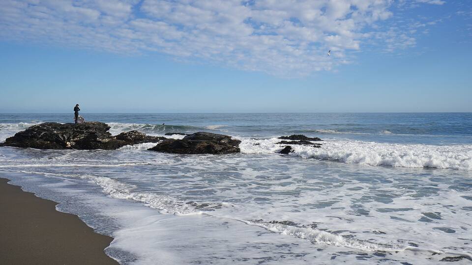A beach with waves, a person on a rock, and a clear sky.
