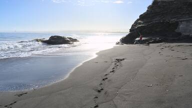A beach with footprints, waves, and a rocky cliff with a railing.