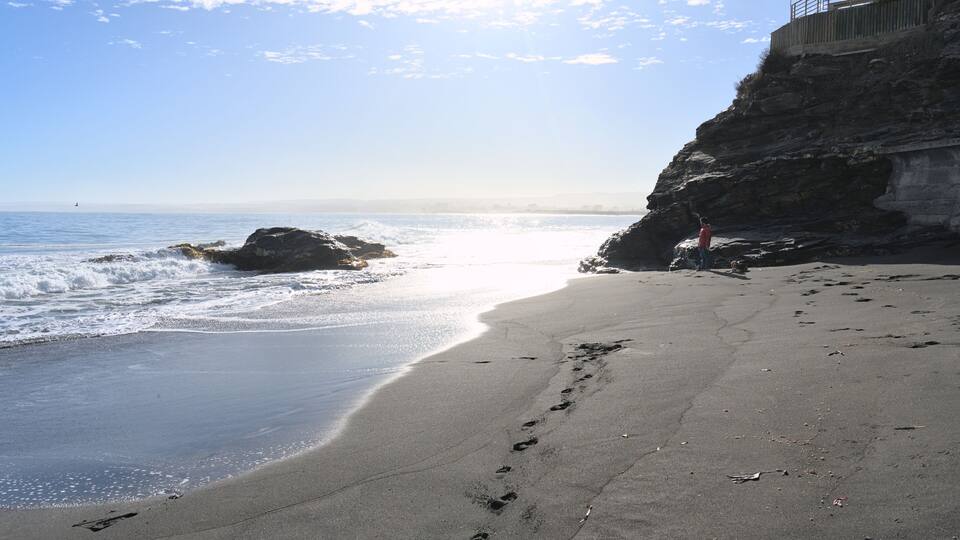 A beach with footprints, waves, and a rocky cliff with a railing.