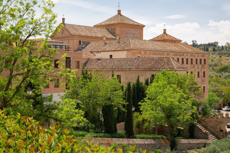 Un bâtiment historique, doté d’un toit recouvert de tuiles, entouré d’une végétation luxuriante et d’arbres.