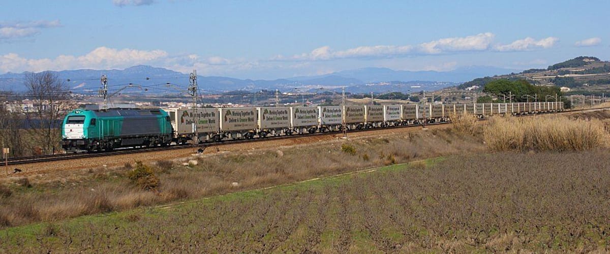 A green train with cargo cars traveling through a rural landscape with fields and distant mountains.