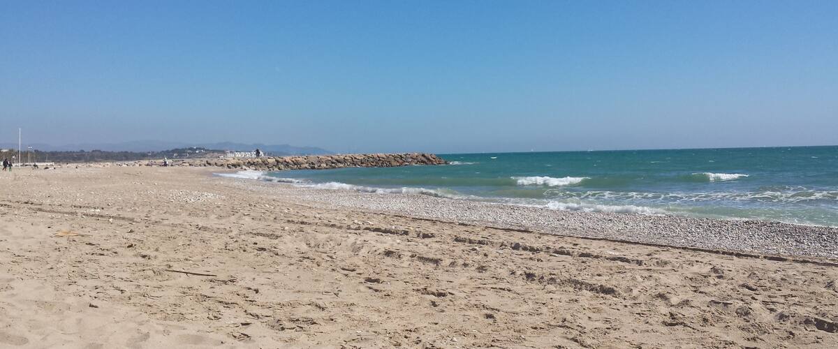 A sandy beach with gentle waves, a rocky breakwater, and a clear blue sky.