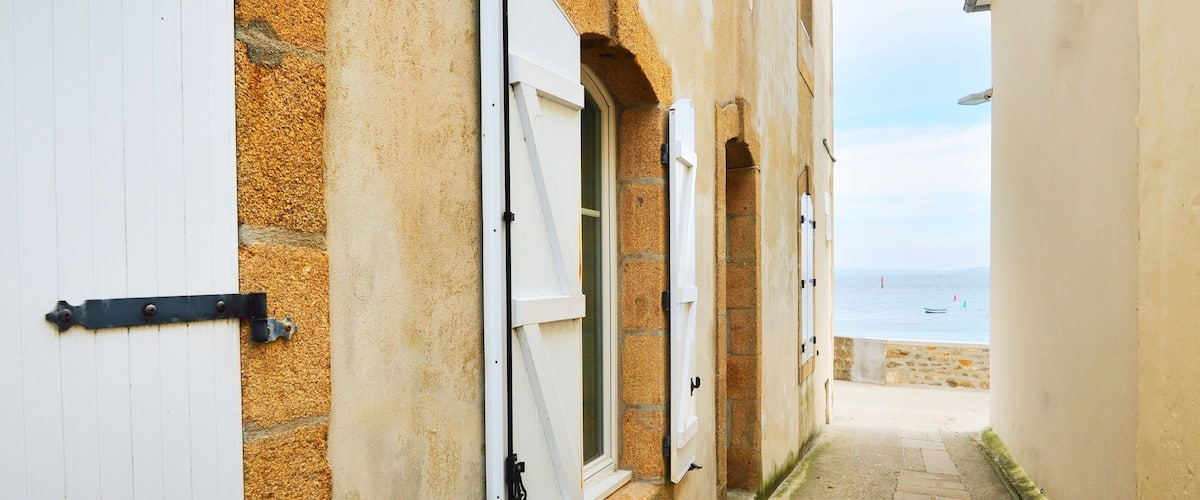 A narrow alleyway with stone walls and white doors, leading to a beach with the sea in the background.
