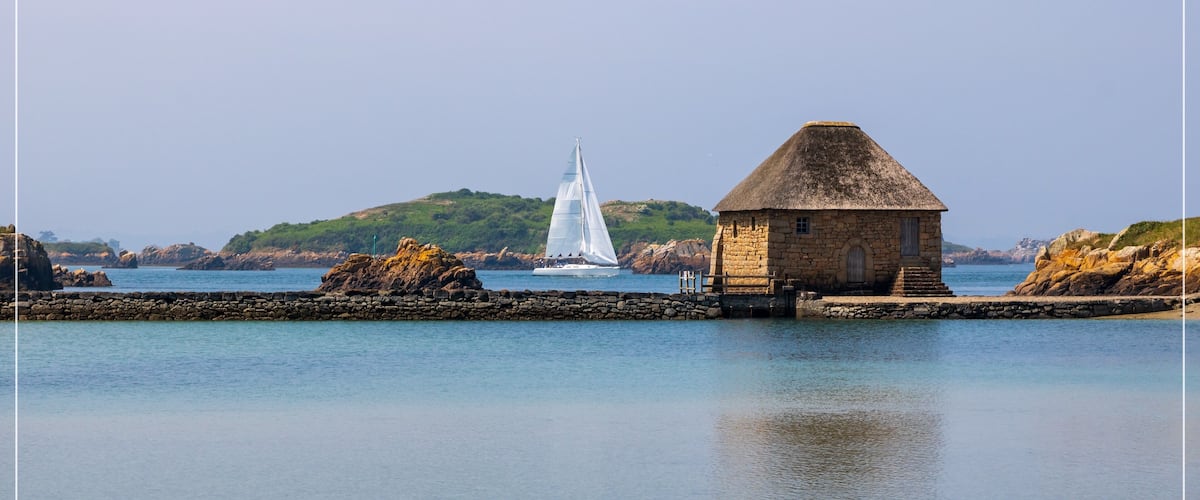 A stone building with a thatched roof near calm waters, a sailboat in the distance, and green hills in the background.