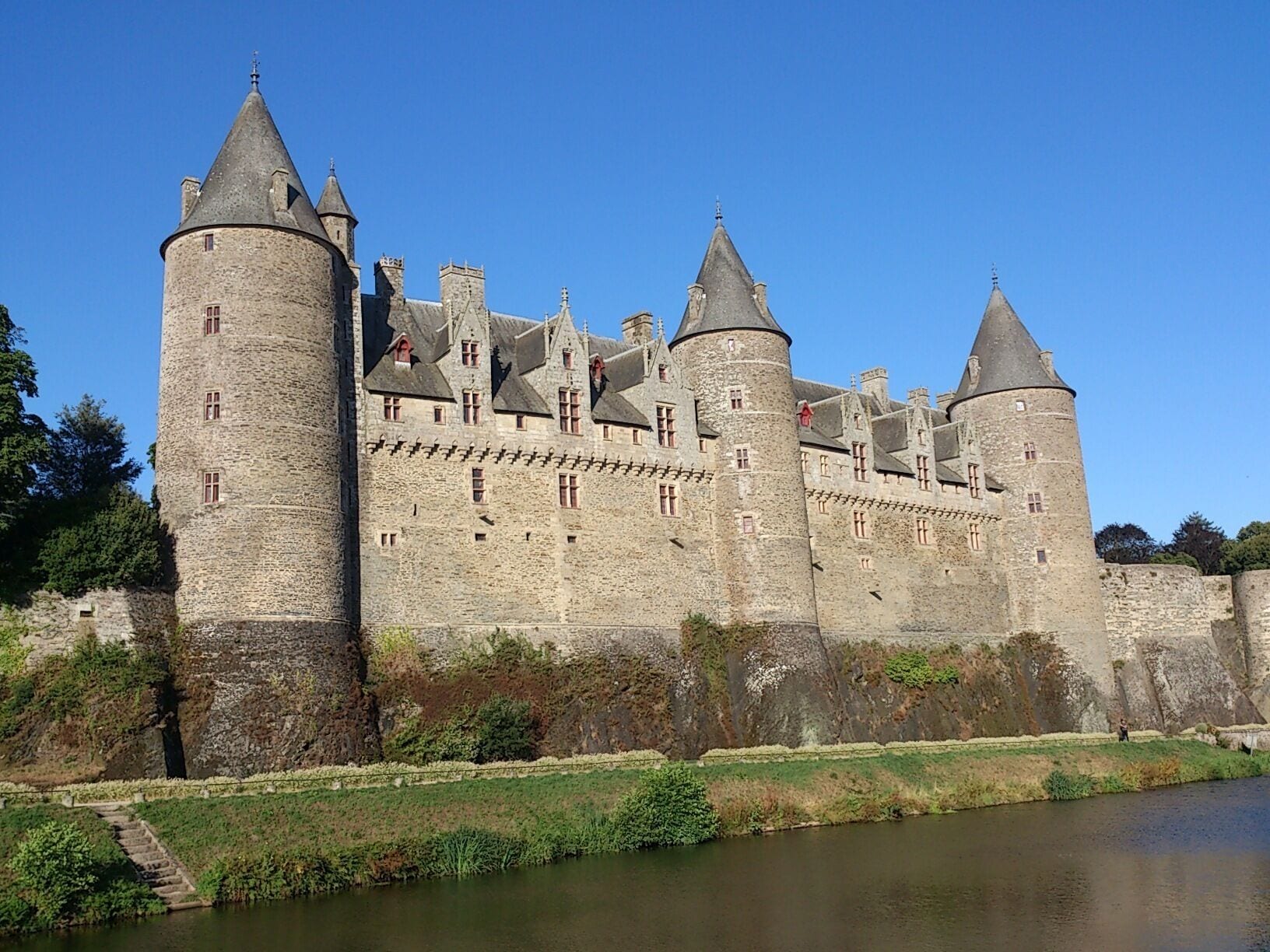 A historic castle with multiple towers, a moat, and a river in the foreground.