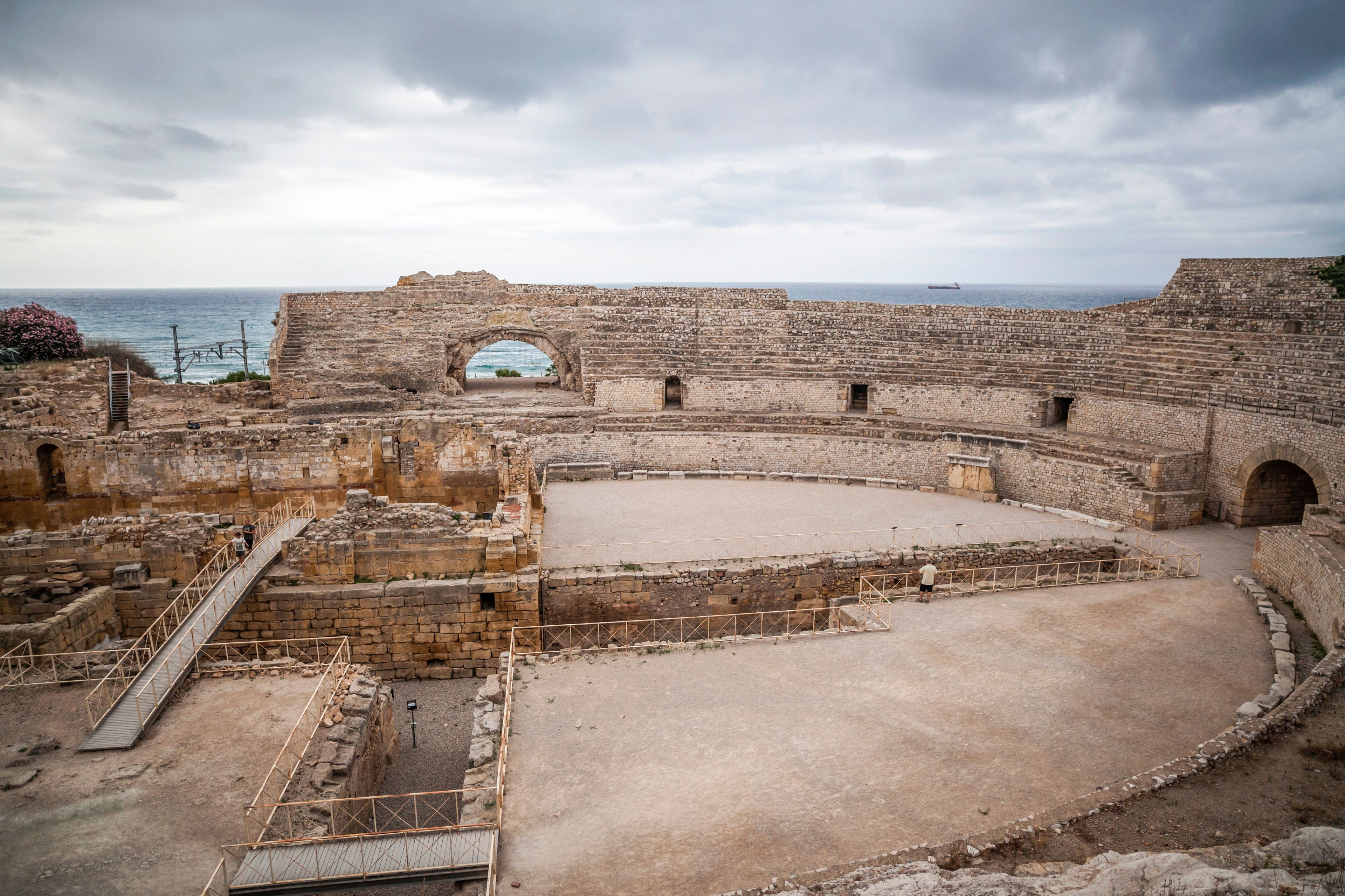 An ancient amphitheater with stone steps and an open central area, overlooking the sea.