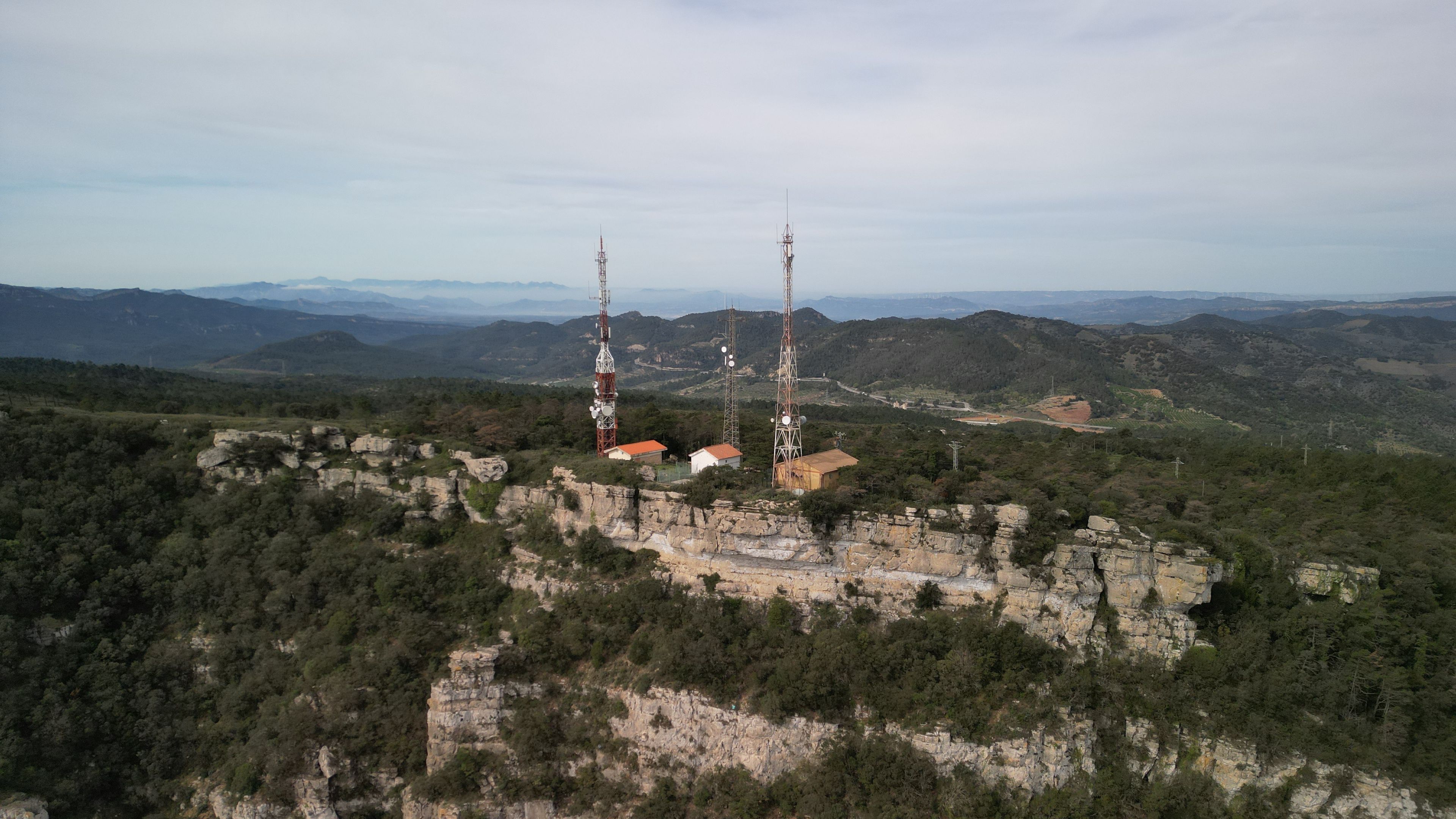 A mountainous landscape with two tall communication towers, a few buildings, and a valley in the background.