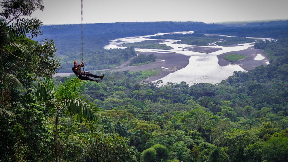 A person bungee jumping from a high cliff over a lush green forest with a river below.