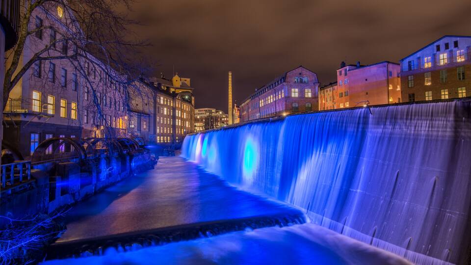 Vista nocturna de una ciudad con una gran cascada iluminada, edificios históricos y una torre destacada.