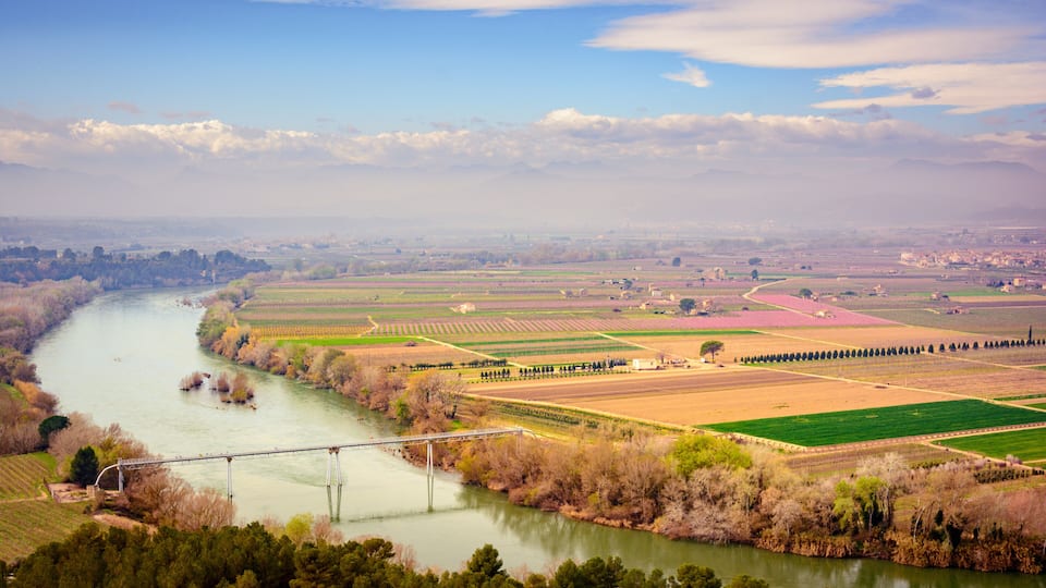 A river winding through agricultural fields, a bridge, and distant mountains under a partly cloudy sky.