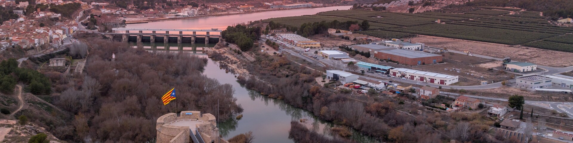 Un atardecer sobre un río, con un puente, una estructura similar a un castillo y edificios industriales.